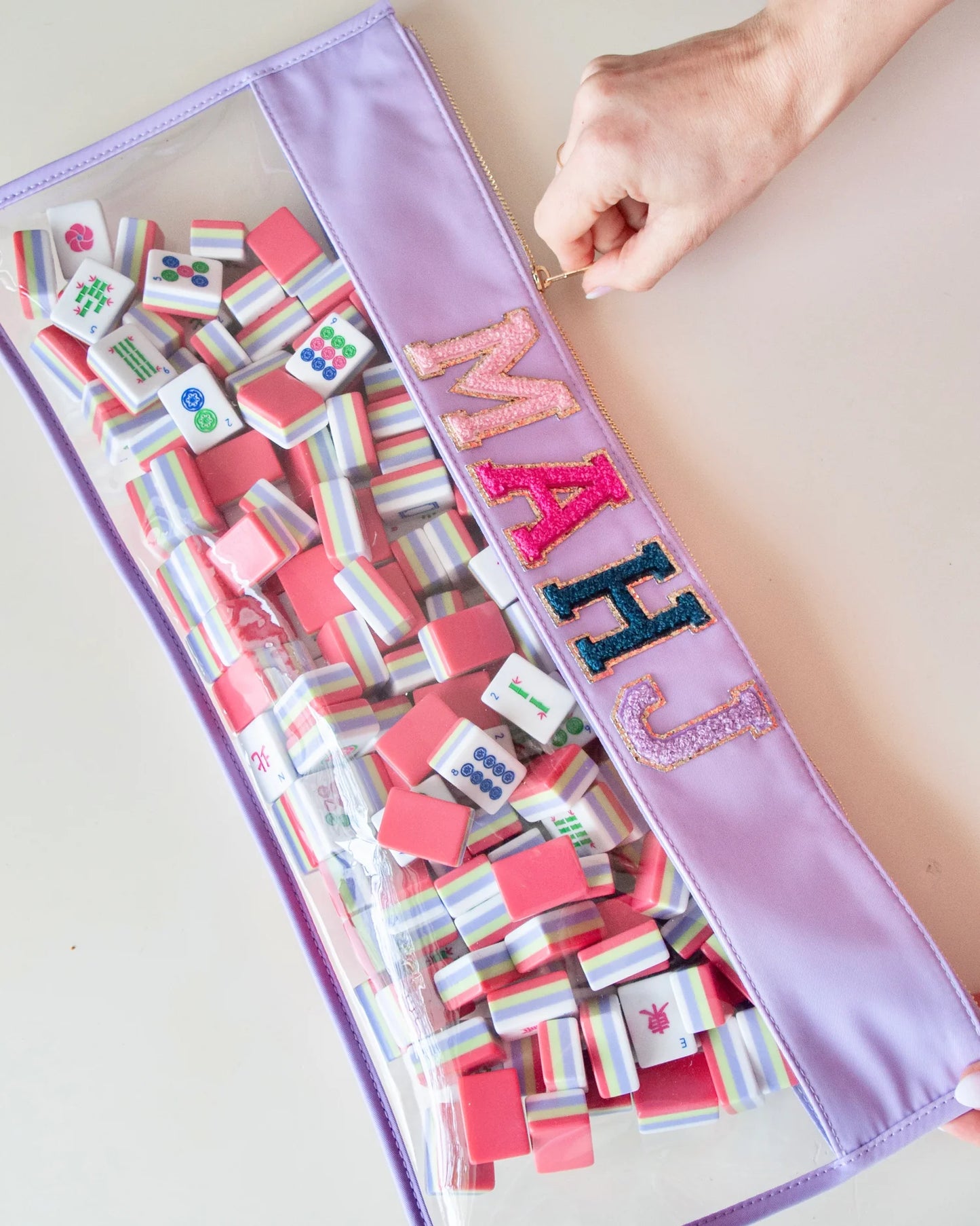 A person's hand unzipping a purple and clear zippered bag  with the letters MAHJ in light pink, hot pink,  blue and purple filled with mahjong tiles.