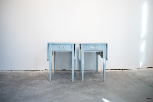 pair of light blue lacquered drop leaf end tables against a white wall on a concrete floor.