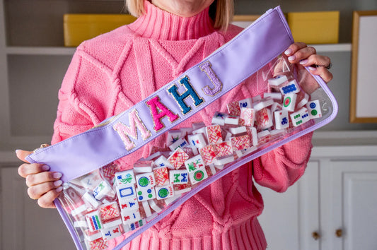 Woman in a pink sweater holding a purple and clear zippered bag  with the letters MAHJ in light pink, hot pink,  blue and purple filled with mahjong tiles.